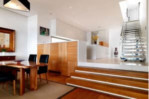 Modern, open-plan dining area with wooden furniture and flooring, leading up steps to a white kitchen and floating staircase with metal handrails.