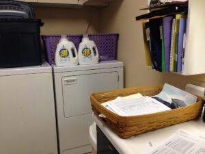 A laundry room with a washer, dryer, bottles of All detergent, a basket of papers, files organized on a wall shelf, and hidden confessions tucked among the paperwork.