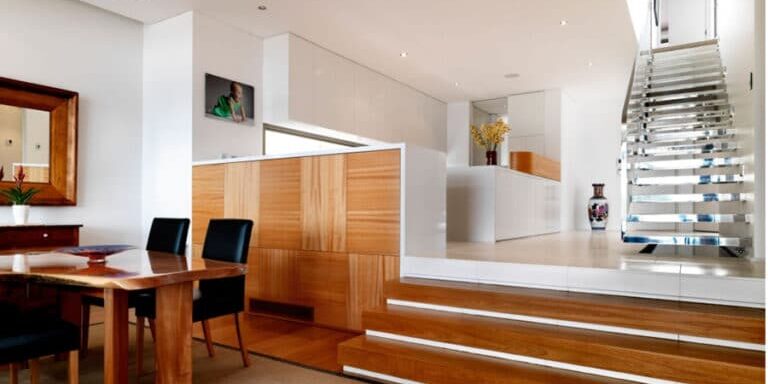 Modern, open-plan dining area with wooden furniture and flooring, leading up steps to a white kitchen and floating staircase with metal handrails.