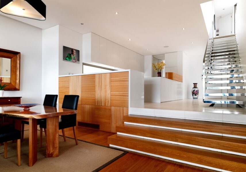 Modern, open-plan dining area with wooden furniture and flooring, leading up steps to a white kitchen and floating staircase with metal handrails.