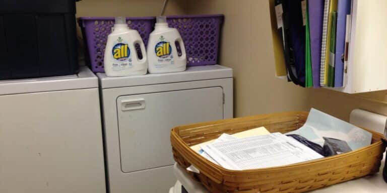 A laundry room with a washer, dryer, bottles of All detergent, a basket of papers, files organized on a wall shelf, and hidden confessions tucked among the paperwork.