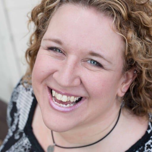 A woman with curly blonde hair, wearing a black and white patterned top and a necklace, smiles broadly at the camera—embodying the confidence found in Portland interior design for women.