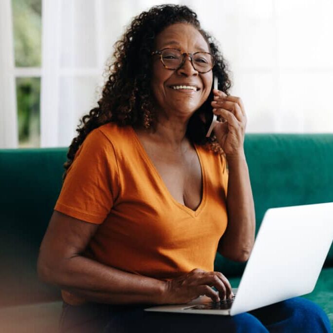 An older woman sits on a green couch, smiling while talking on a smartphone and using a laptop computer, enjoying stylish interior design for women Portland is known for.